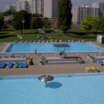 Öffentlicher Freibadkomplex mit mehreren Becken, Liegestühlen und Sonnenschirmen, umgeben von Gras und Wohnhäusern im Hintergrund.