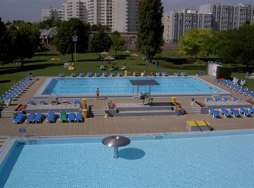 Öffentlicher Freibadkomplex mit mehreren Becken, Liegestühlen und Sonnenschirmen, umgeben von Gras und Wohnhäusern im Hintergrund.