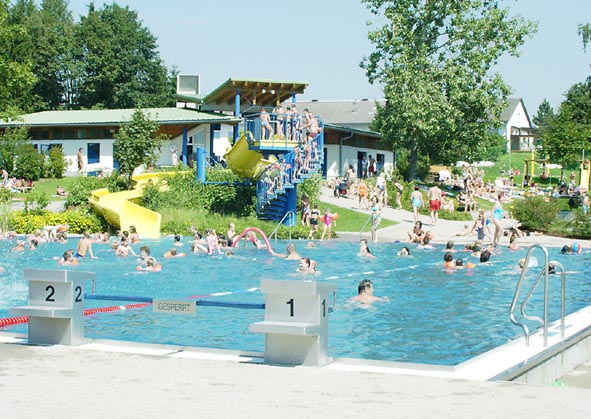 Öffentliches Freibad mit vielen Menschen, die schwimmen und sich entspannen; an einem sonnigen Tag sind im Hintergrund eine gelbe Rutsche und Gebäude zu sehen.