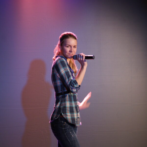 Eine Person in einem karierten Hemd hält ein Mikrofon in der Hand und performt auf der Bühne unter rosa und blauer Beleuchtung.