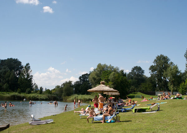 An einem sonnigen Tag entspannen sich die Menschen auf Liegestühlen und schwimmen in einem kleinen, von Bäumen umgebenen See, der von einem strohgedeckten Sonnenschirm beschattet wird.
