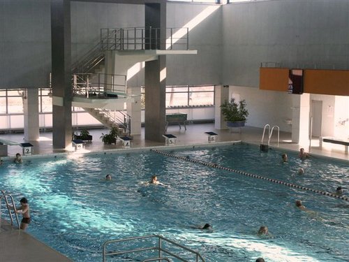 Hallenbad mit mehreren schwimmenden Personen und einem großen Sprungturm im Hintergrund. Das Sonnenlicht strömt durch die oberen Fenster ein und erhellt den Raum.