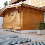 Eine kleine Holzhütte mit schrägem Dach steht neben Bänken, Topfpflanzen und gestreiften Liegestühlen in der Nähe eines Swimmingpools.