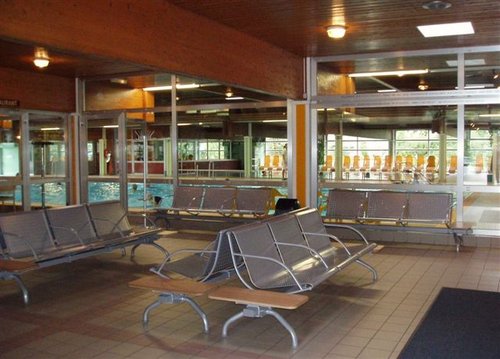 Innenwartebereich mit Metallbänken vor großen Fenstern mit Blick auf ein Schwimmbad. Der Bereich hat geflieste Böden und eine Holzdecke mit Oberlichtern.
