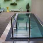 Kleiner Innenpool mit Metallgeländern, umgeben von Liegestühlen und Topfpflanzen vor einer grünen Wand mit hängenden Pflanzgefäßen.