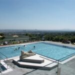Freibad mit einer Statue auf einer Plattform in der Mitte, mehreren schwimmenden Personen und einem Blick auf die Stadt im Hintergrund unter einem klaren Himmel.