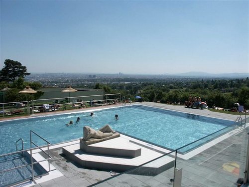 Freibad mit einer Statue auf einer Plattform in der Mitte, mehreren schwimmenden Personen und einem Blick auf die Stadt im Hintergrund unter einem klaren Himmel.