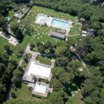 Luftaufnahme eines Freibadkomplexes mit mehreren Becken, umliegenden grünen Rasenflächen, verstreuten Liegestühlen und Bäumen.