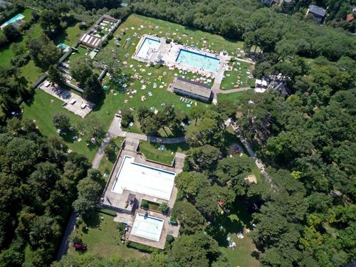 Luftaufnahme eines Freibadkomplexes mit mehreren Becken, umliegenden grünen Rasenflächen, verstreuten Liegestühlen und Bäumen.