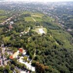 Luftaufnahme einer grünen, hügeligen Landschaft mit verstreuten Gebäuden, Parkplätzen und Bäumen, die sich unter klarem Himmel in Richtung einer entfernten Stadtlandschaft erstreckt.