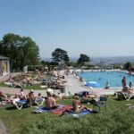 Menschen sonnen sich und schwimmen in einem überfüllten Freibad an einem sonnigen Tag, während im Hintergrund die Stadt zu sehen ist.