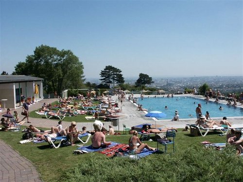 Menschen sonnen sich und schwimmen in einem überfüllten Freibad an einem sonnigen Tag, während im Hintergrund die Stadt zu sehen ist.