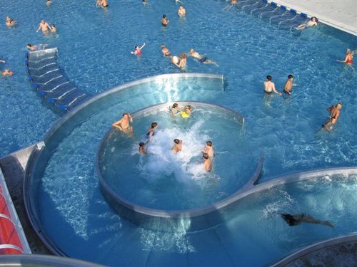 Luftaufnahme eines überfüllten Freibads mit schwimmenden, watenden und spielenden Menschen in einem kreisförmigen Beckenbereich, der mit dem Hauptbecken verbunden ist.