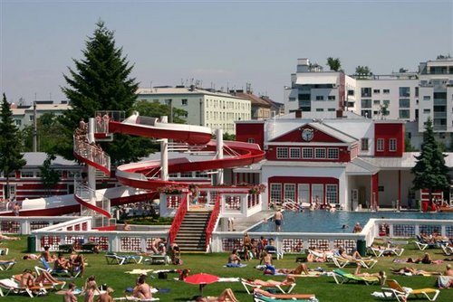 Die Menschen entspannen sich auf Liegestühlen und im Gras in der Nähe eines Außenpools mit einer rot-weißen Spiralwasserrutsche, umgeben von Gebäuden und Bäumen.