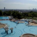 Öffentlicher Freibadkomplex mit mehreren Becken, schwimmenden und faulenzenden Menschen und einer im Hintergrund sichtbaren Stadtsilhouette bei teilweise bewölktem Himmel.