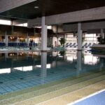 Hallenbad mit klarem Wasser, gefliestem Boden, Liegestühlen an der Seite und großen Fenstern, die viel Tageslicht hereinlassen.