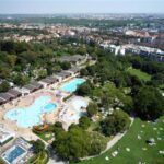 Luftaufnahme eines großen öffentlichen Freibadkomplexes mit mehreren Becken, umgeben von Grünanlagen und Gebäuden im Hintergrund.