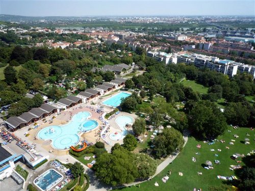 Luftaufnahme eines großen öffentlichen Freibadkomplexes mit mehreren Becken, umgeben von Grünanlagen und Gebäuden im Hintergrund.