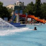 Freibad mit Menschen, einem Wasserspiel im Vordergrund und einer orangefarbenen Wasserrutsche im Hintergrund in der Nähe von Bäumen und Gebäuden.