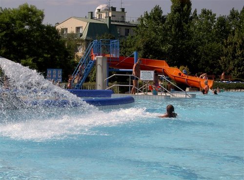 Freibad mit Menschen, einem Wasserspiel im Vordergrund und einer orangefarbenen Wasserrutsche im Hintergrund in der Nähe von Bäumen und Gebäuden.