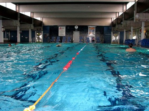 Hallenbad mit Bahnen, die durch ein schwimmendes Seil getrennt sind, ein paar Schwimmer im Wasser und Liegestühle an den Wänden.