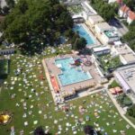 Luftaufnahme eines öffentlichen Freibadkomplexes mit Menschen, die in einer parkähnlichen, von Bäumen und Gebäuden umgebenen Umgebung schwimmen, sich auf dem Rasen räkeln und auf Handtüchern sonnen.