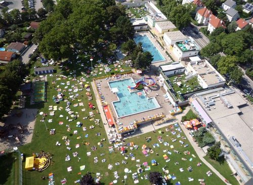 Luftaufnahme eines öffentlichen Freibadkomplexes mit Menschen, die in einer parkähnlichen, von Bäumen und Gebäuden umgebenen Umgebung schwimmen, sich auf dem Rasen räkeln und auf Handtüchern sonnen.