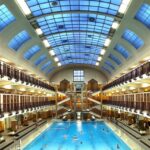 Hallenbad mit Personenschwimmen, umgeben von zwei Ebenen mit Spinden und Bänken und einer großen gewölbten Glasdecke, die natürliches Licht hereinlässt.