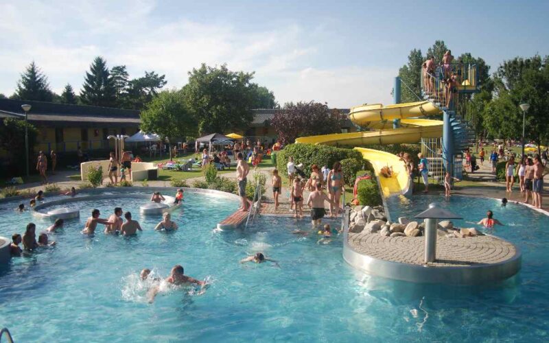 Öffentliches Freibad mit Menschen, die an einem sonnigen Tag schwimmen, faulenzen und darauf warten, die gelben Wasserrutschen zu benutzen. Bäume und Rasenflächen umgeben den Poolbereich.