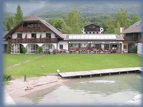 Ein großes Haus am See mit grünem Rasen, Sitzgelegenheiten im Freien und einem hölzernen Steg vor der Kulisse von Bäumen und Bergen.