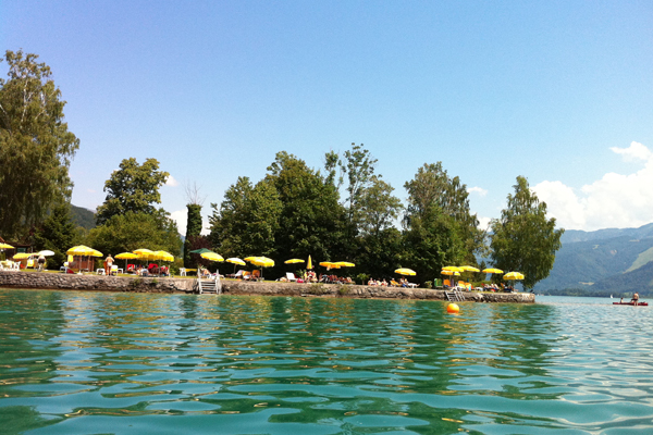 Blick auf ein Seeufer mit gelben Sonnenschirmen, Bäumen und Menschen, die sich am Wasser entspannen; im Hintergrund sind bei klarem Himmel die Berge zu sehen.