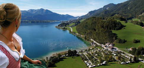 Eine Frau in traditioneller Kleidung blickt auf einen See, umgeben von grünen Hügeln, Bergen und einer kleinen Stadt unter einem klaren blauen Himmel.