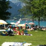 Menschen, die sich auf einer Wiese an einem See sonnen und entspannen, mit Sonnenschirmen und Handtüchern; im Hintergrund sind Berge und eine Kirche zu sehen.