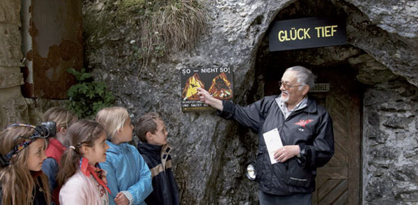 Ein älterer Mann erklärt einer Gruppe von Kindern die Sicherheitszeichen am Eingang einer Höhle mit der Aufschrift "Glück Tief".