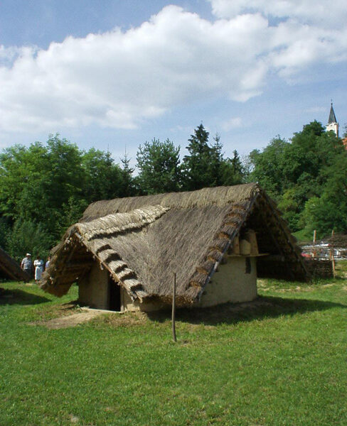 Eine kleine, strohgedeckte Hütte steht auf einer Wiese mit Bäumen und einem Kirchturm im Hintergrund unter einem teilweise bewölkten Himmel.