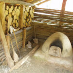 Ein rustikaler, mit Holz befeuerter Lehmofen steht unter einem hölzernen Unterstand, an dessen Wand gestapeltes Brennholz und große hölzerne Backwerkzeuge lehnen.