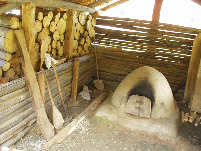Ein rustikaler, mit Holz befeuerter Lehmofen steht unter einem hölzernen Unterstand, an dessen Wand gestapeltes Brennholz und große hölzerne Backwerkzeuge lehnen.