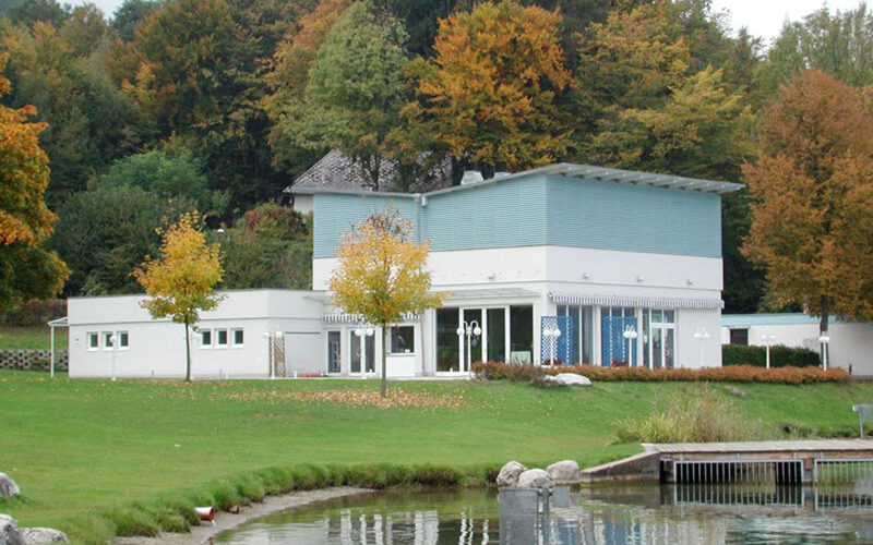 Modernes weiß-blaues Gebäude mit großen Fenstern, umgeben von Bäumen mit Herbstlaub, neben einer Wiese und einem Holzsteg am Wasser.