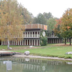 Ein modernes, zweistöckiges Gebäude mit vielen vertikalen Fenstern steht an einem Teich, umgeben von grünem Gras, Bäumen und etwas Herbstlaub.
