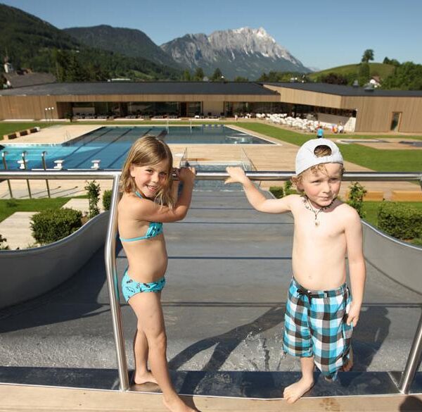 Zwei kleine Kinder in Badekleidung stehen an der Spitze einer Wasserrutsche in einem Freibadkomplex mit Bergen im Hintergrund.