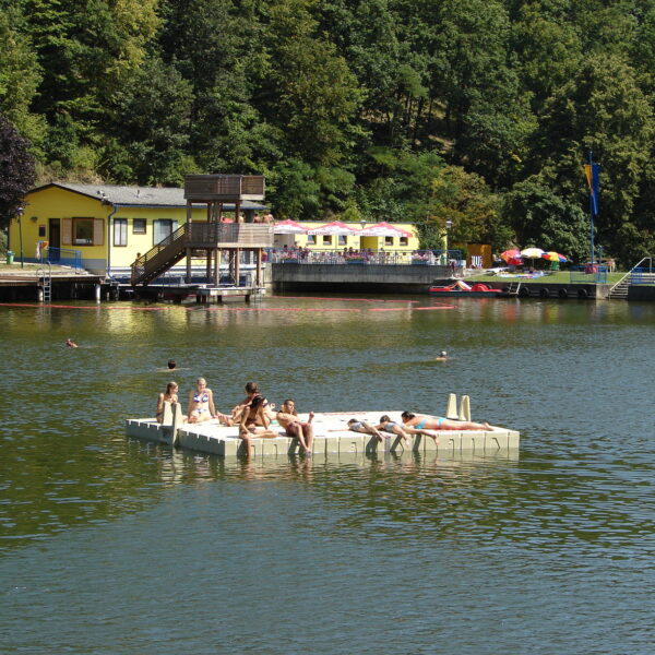 Menschen entspannen sich auf einer schwimmenden Plattform in einem See mit einem Gebäude und Bäumen im Hintergrund an einem sonnigen Tag.