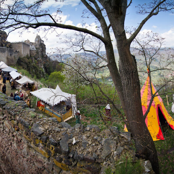 Ein mittelalterlicher Markt mit Zelten und Menschen ist entlang der Steinmauern einer alten Burg auf einem Hügel aufgebaut, umgeben von Bäumen und fernen Bergen.