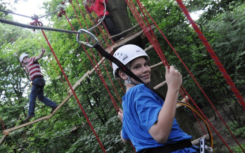 Kinder, die Helme und Klettergurte tragen, nehmen an einem Hochseilgarten im Freien teil und klettern und balancieren auf erhöhten Plattformen zwischen Bäumen.