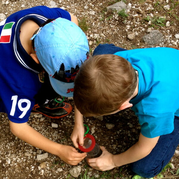 Zwei Jungen kauern auf felsigem Boden und untersuchen etwas in einem roten Vergrößerungsglas oder einem Käferbehälter.