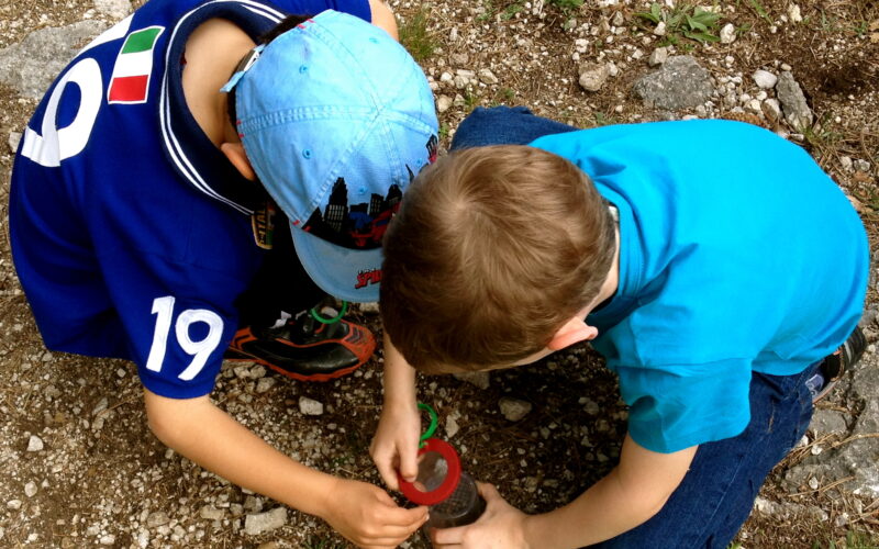 Zwei Jungen kauern auf felsigem Boden und untersuchen etwas in einem roten Vergrößerungsglas oder einem Käferbehälter.