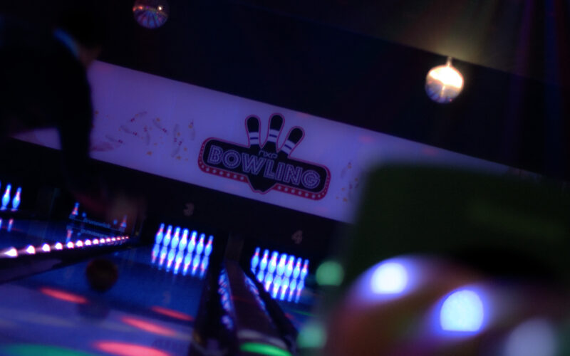 Eine leuchtende Bowlingbahn mit Neonlicht und einem Schild mit der Aufschrift "Bowling" über den Bahnen; am Ende der Bahnen sind Bowlingpins beleuchtet.