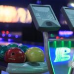 Nahaufnahme einer Bowlingkugel-Rückgabe mit bunten Kugeln und zwei elektronischen Scoring-Bildschirmen in einer Bowlingbahn.