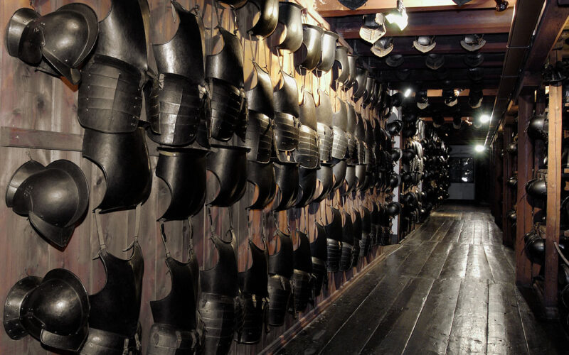 An den Holzwänden entlang eines schwach beleuchteten Korridors mit Holzboden sind Reihen von schwarzen Metallpanzern und Helmen angebracht.