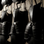 An einer Holzwand hängen reihenweise mittelalterliche Plattenrüstungen, wobei die Schatten die dunklen Metalloberflächen hervorheben.