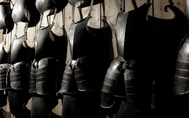 An einer Holzwand hängen reihenweise mittelalterliche Plattenrüstungen, wobei die Schatten die dunklen Metalloberflächen hervorheben.